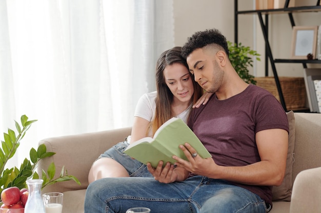 Lindo casal jovem sentado no sofá em casa lendo um livro interessante juntos