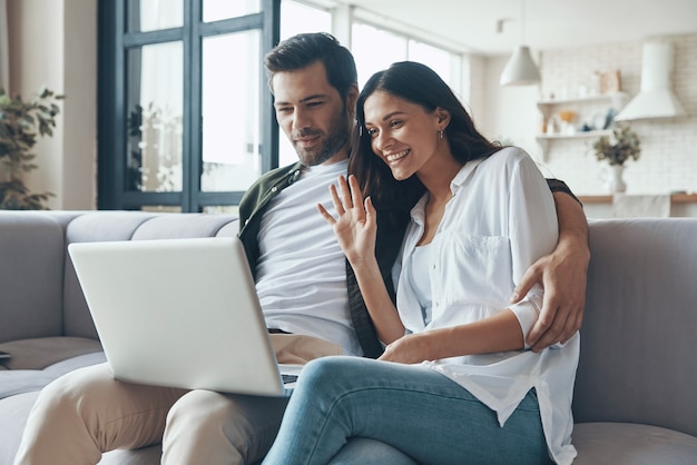 Lindo casal jovem tomando café e olhando para o laptop enquanto está sentado no sofá em casa