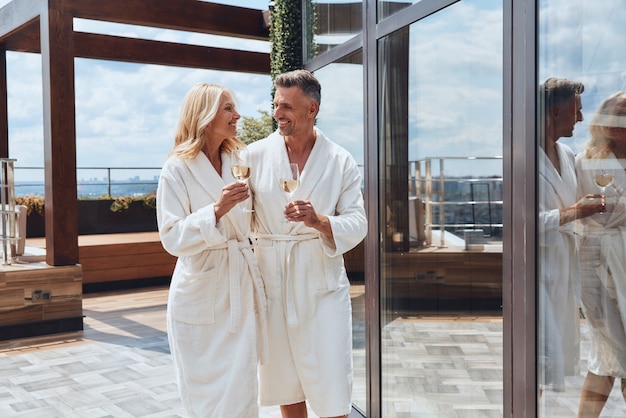 Foto lindo casal maduro em roupões de banho, desfrutando de champanhe enquanto relaxa em um hotel de luxo ao ar livre