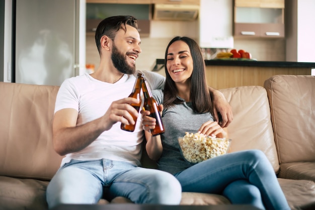 Lindo casal moderno e sorridente apaixonado está assistindo a algum filme ou filme na TV enquanto relaxa no sofá do próprio apartamento