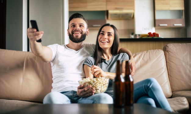 Lindo casal moderno e sorridente apaixonado está assistindo a algum filme ou filme na TV enquanto relaxa no sofá do próprio apartamento