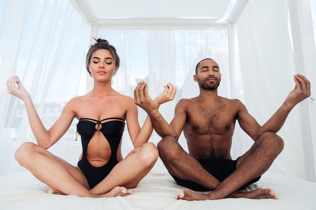 Lindo casal multirracial meditando e sentado em posição de lótus na cama da praia