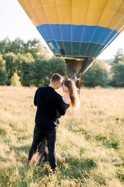 Lindo casal romântico em roupas pretas, abraçando e dançando em um lindo prado verde de verão com um balão de ar quente amarelo