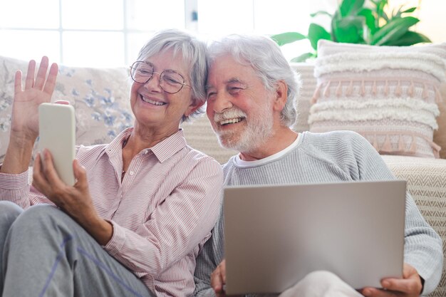 Lindo casal sênior caucasiano sentado no chão em casa usando laptop e telefone celular idosos aposentados modernos usando dispositivos tecnológicos