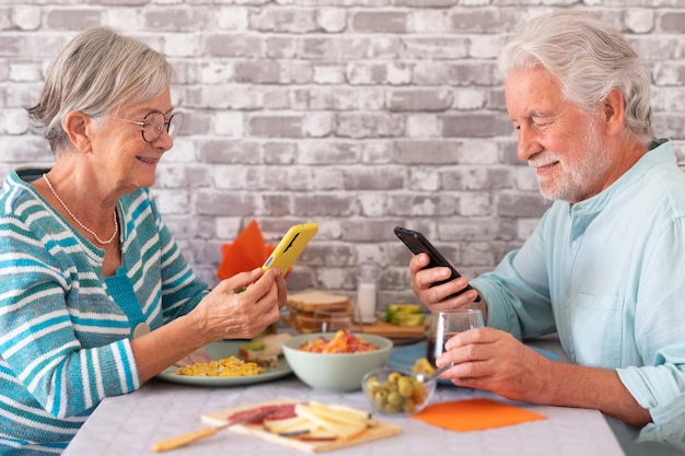 Lindo casal sênior sentado cara a cara na mesa usando seus telefones celulares enquanto brunch juntos em casa