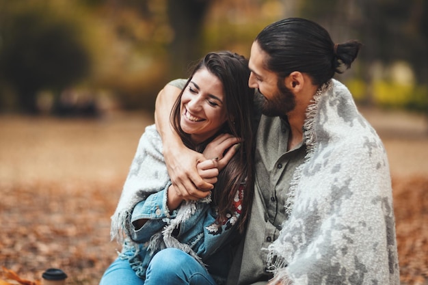 Lindo casal sorridente desfrutando no parque da cidade ensolarada em cores de outono, olhando um ao outro. Eles estão sentados no chão e se divertindo.