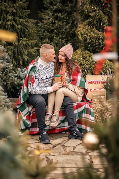 Lindo casal sorridente feliz embrulhado em um cobertor escocês Árvores de Natal verdes ao fundo As férias de inverno Esperando o ano novo