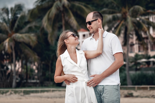 Lindo casal vestido de branco abraçando na praia na Tailândia; palmeiras e hotel ao fundo; conceito de aniversário.