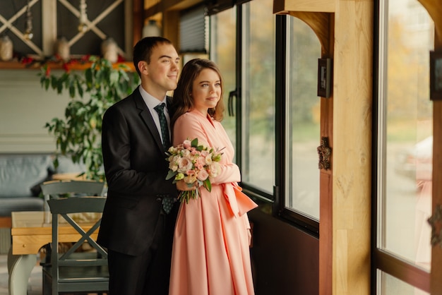 Lindo casamento, marido e mulher, noivos em pé no interior do sotão perto da janela. recém-casados casal apaixonado. noivo abraça a noiva pelos ombros. noivo segurar um buquê de casamento nas mãos.