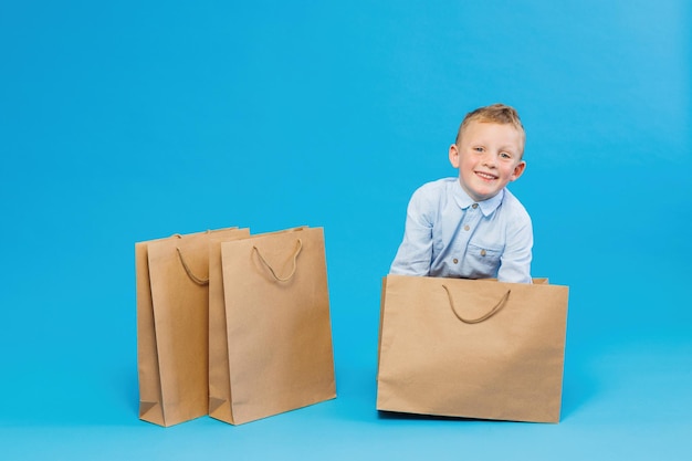 Lindo chico divertido con camisa azul sentado cerca de bolsas de papel aisladas en fondo azul en el estudio Concepto de estilo de vida de la gente El chico se ofrece a ir de compras