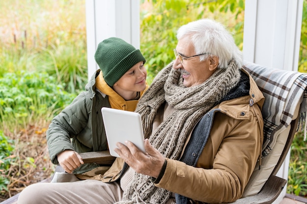 Lindo colegial sonriente en gorro y chaqueta abrigada mirando a su abuelo con tableta digital mientras habla de noticias al aire libre