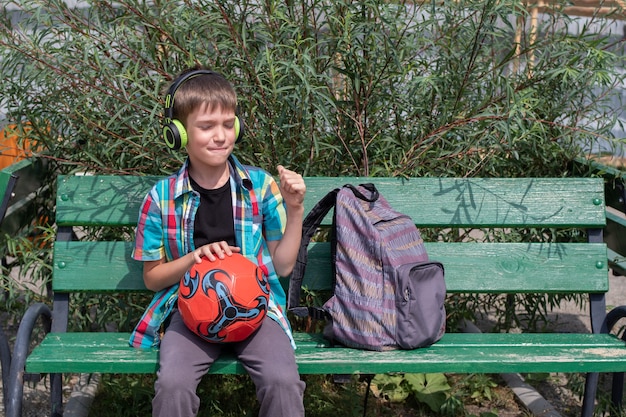 lindo colegial vestido con ropa casual escucha música con auriculares, se sienta en un banco con un balón de fútbol. Concepto de regreso a la escuela