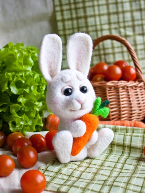 Lindo conejito blanco de fieltro de aguja con zanahoria en las manos en el fondo de verduras