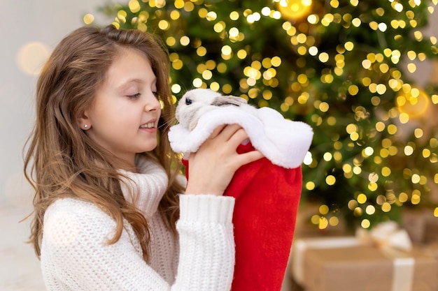 Foto lindo conejito de navidad símbolo del año nuevo 2022 primer plano en el sombrero de santa en manos de un niño feliz en el fondo de un árbol de navidad decorado con luces de desenfoque