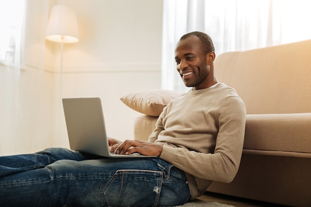 Lindo día. Hombre afroamericano con barba exuberante sonriendo y trabajando en la computadora portátil y escribiendo mientras está sentado en el piso cerca del sofá