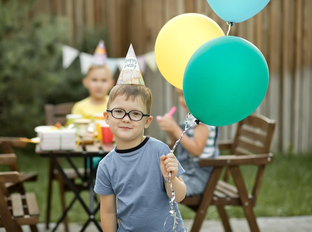 Lindo y divertido niño de cuatro años celebrando su cumpleaños con familiares o amigos en una fiesta de cumpleaños en el patio trasero Niño con sombrero de fiesta y sosteniendo un globo en una mano