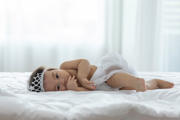 Lindo dulce adorable bebé asiático con vestido de novia y diadema sentado en la cama blanca sonriendo y jugando con felicidad emocional en un dormitorio acogedor, concepto de bebé saludable