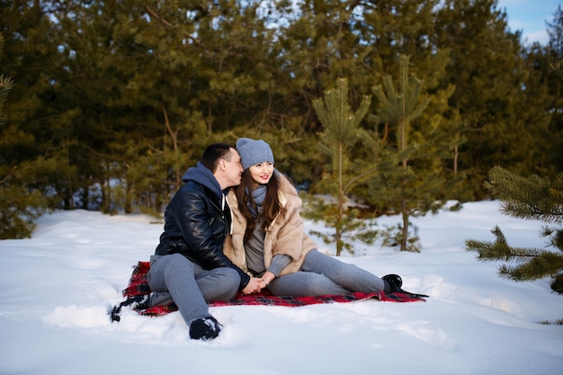 Lindo e feliz casal apaixonado, sentado em um cobertor no inverno em um bosque nevado