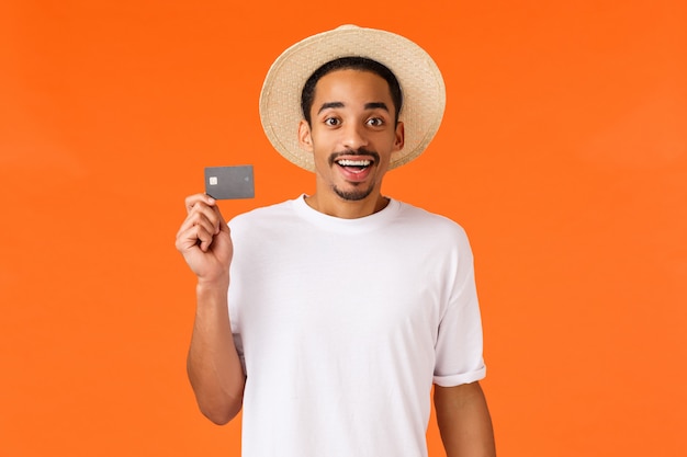 Lindo y emocionado hombre afroamericano compró vacaciones de viaje para vacaciones con dinero de crédito, con tarjeta y sonriendo divertido, siendo cliente premium banco sentado aeropuerto vip terminal, pared naranja