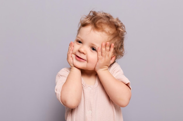 Lindo encantador bebé sonriente bebé niña caucásica posando aislada sobre fondo gris mirando hacia otro lado con una sonrisa tocándose la cara ve algo interesante