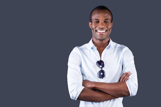 Lindo encantador. Belo jovem negro com camisa azul, de braços cruzados e sorrindo para a câmera, em pé contra um fundo cinza