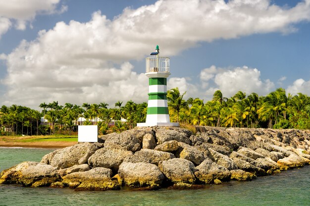 Lindo farol com listras no porto pedregoso perto da água, dia ensolarado ao ar livre com palmeiras verdes e céu azul nublado no verão