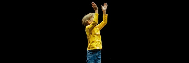 Foto lindo, feliz, sincero, caucásico, niño pequeño de seis años con cabello largo y rubio, se para alto y salta y aplaude en el estudio iluminado por una pancarta de luz blanca brillante amarilla