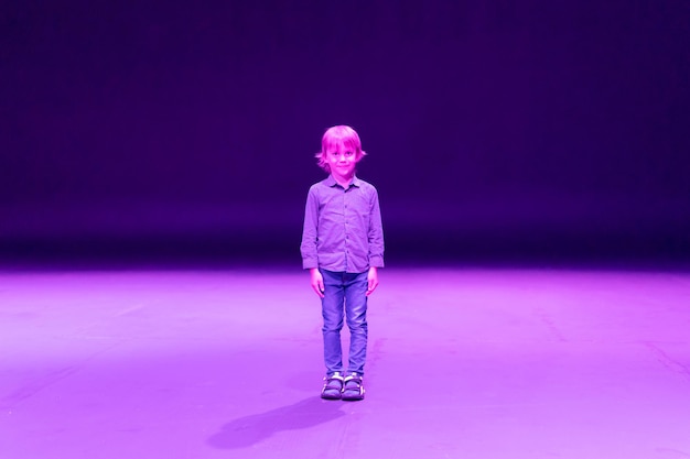 Lindo, feliz, sincero, caucásico, niño pequeño de seis años con cabello largo y rubio, se encuentra alto en el estudio iluminado por luz de neón púrpura viva magenta