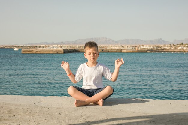 Lindo, feliz, sonriente, pequeño, niño, niño, niño, con, ojos cerrados, prácticas, yoga, meditar, aire libre, en, mar