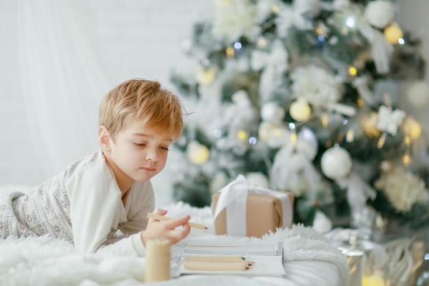 Lindo garotinho de pijama com estrelas comemora o natal. feriados de ano novo. as crianças brincam no trenó de natal branco na sala festivamente decorada, copie o espaço na área direita.