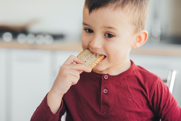 Lindo garotinho na cozinha comendo um pedaço de pão com fome