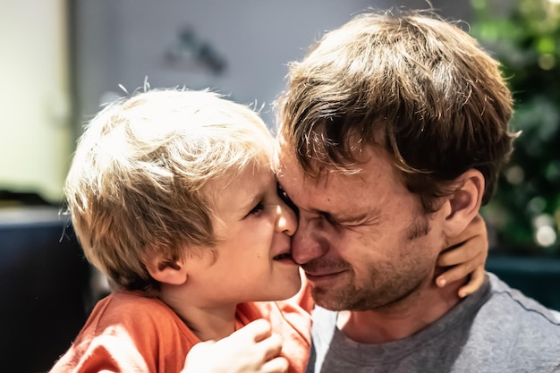 Lindo hijo pequeño niño rubio abraza suavemente al padre por el cuello ojos cerrados con placer divertido travieso cara a cabeza dentro de la habitación Infancia feliz paternidad sentimientos de amor día del padre piel real