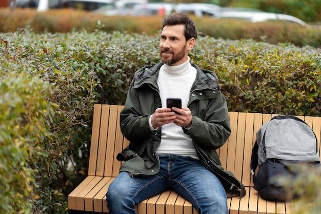 Lindo hombre sin afeitar está sentado en un banco sosteniendo un teléfono en sus manos El chico mira hacia el lado al aire libre