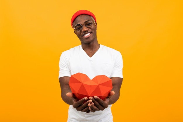 Lindo hombre africano guapo en una camiseta blanca sostiene un corazón rojo 3d hecho de papel para el día de San Valentín en un fondo amarillo