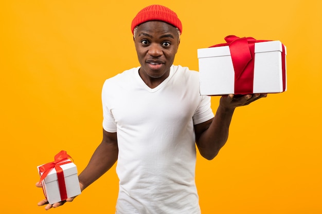 Lindo hombre africano negro sorprendido con una sonrisa en una camiseta blanca tiene dos cajas de regalo con una cinta roja para el día de San Valentín amarillo