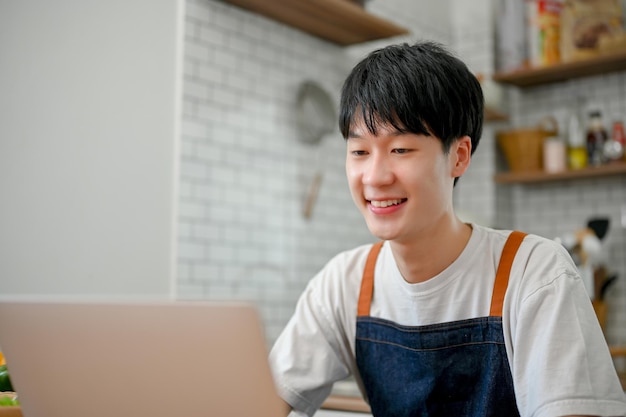 Lindo hombre asiático en delantal usando una computadora portátil en su cocina buscando recetas de comida en Internet