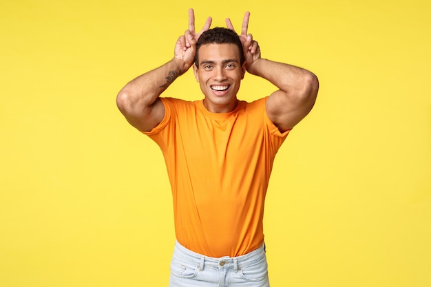 Lindo hombre masculino en camiseta naranja hace orejas de conejo detrás de la cabeza