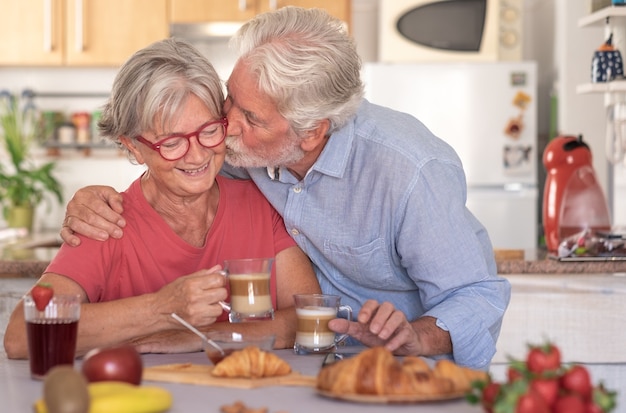 Lindo homem sênior beijando sua esposa tomando café da manhã em casa. Aposentados felizes bebendo cappuccino comendo frutas e croissant