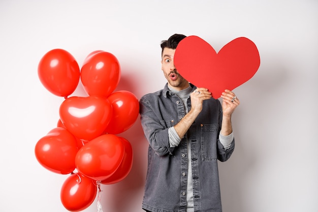 Lindo joven esconde la cara detrás de la gran tarjeta de corazón de San Valentín, admirador secreto mirando a la cámara con asombro, diciendo wow, de pie cerca de globos románticos en blanco.