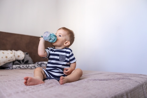 Foto lindo joven rubio sentado en la cama en el dormitorio y bebiendo agua de su botella.