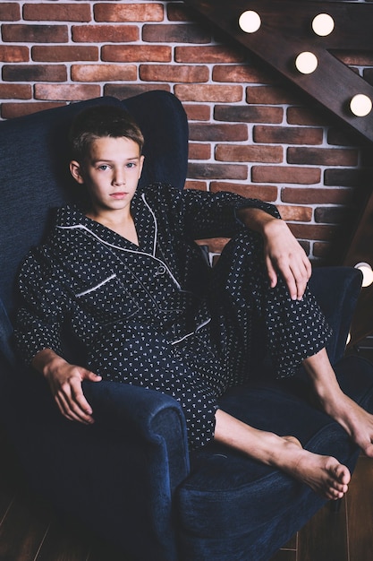 Lindo menino adolescente moderno sentado em uma cadeira em casa de pijama