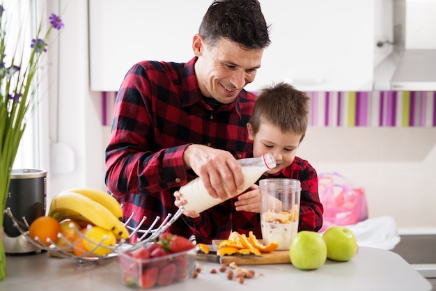 Lindo menino e seu pai amoroso, derramando leite juntos na mistura de frutas na tigela do liquidificador.