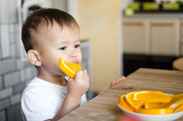 Lindo menino na cozinha comendo uma laranja avidamente cortada em fatias