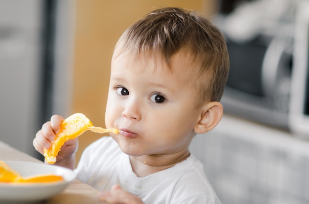 Lindo menino na cozinha comendo uma laranja avidamente cortada em fatias