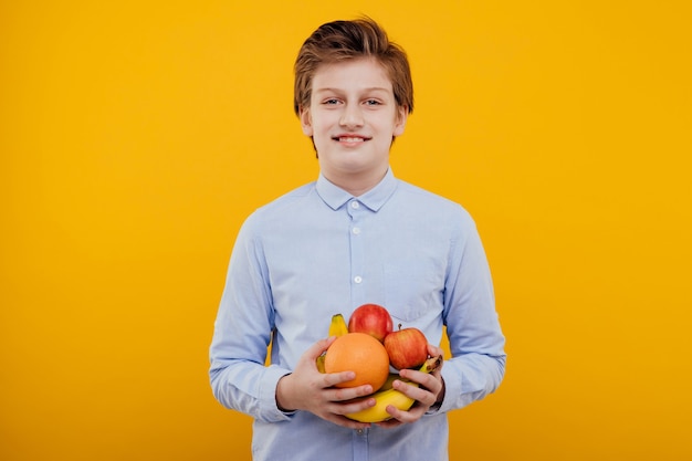 Lindo menino pré-adolescente com frutas na mão, camisa azul, isolado na parede amarela