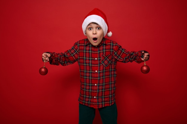 Lindo menino pré-adolescente espantado, criança linda surpresa fica sobre um fundo vermelho com brinquedos brilhantes esféricos de Natal em seus braços, olha para a câmera com surpresa. Conceito de ano novo com espaço de cópia