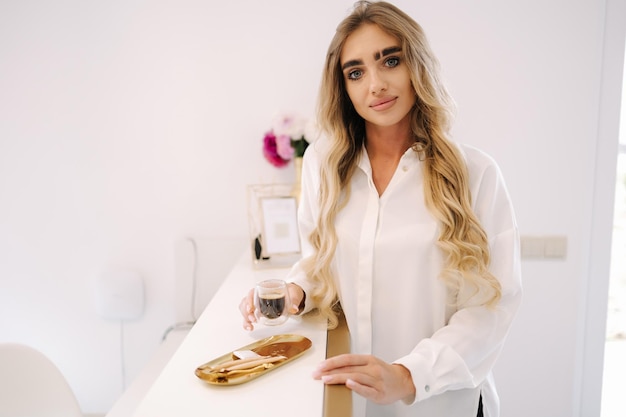 Lindo modelo à espera de procedimento de cosmetologia e beber café no foyer branco interior de beleza