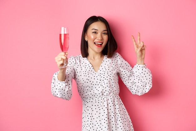 Lindo modelo feminino asiático bebendo champanhe, celebrando na festa e mostrando o símbolo da paz, sorrindo feliz para a câmera, em pé rosa.