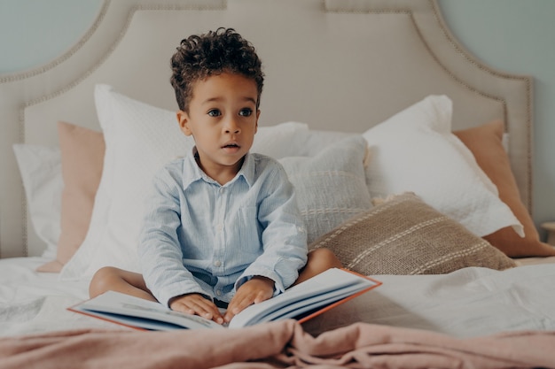Lindo niño afroamericano de pelo rizado sentado en la cama con libro