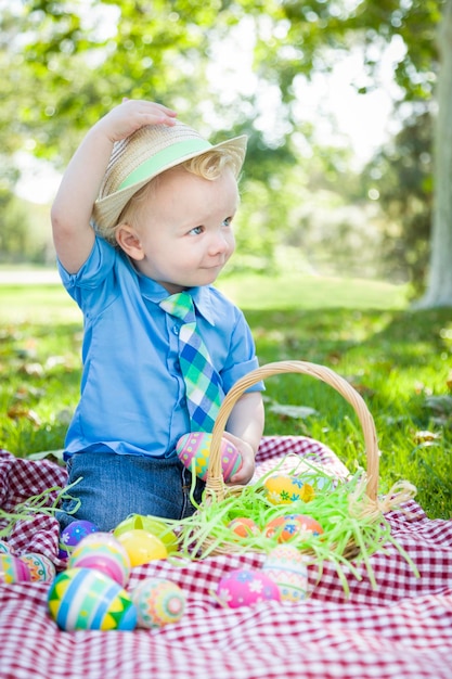 Lindo niño afuera en una manta de picnic sosteniendo huevos de Pascua inclina su sombrero